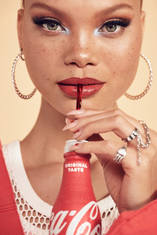 Close-up of a woman drinking Coca-Cola with red lipstick, hoop earrings, and dramatic makeup against a neutral background.
