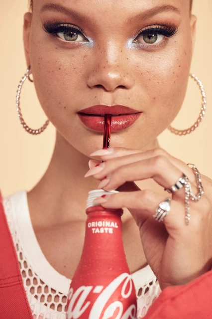 Close-up of a woman drinking Coca-Cola with red lipstick, hoop earrings, and dramatic makeup against a neutral background.