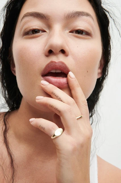 Close-up shot of a woman with fair skin, gold rings, and minimalist tattoos posing in front of a bright background.
