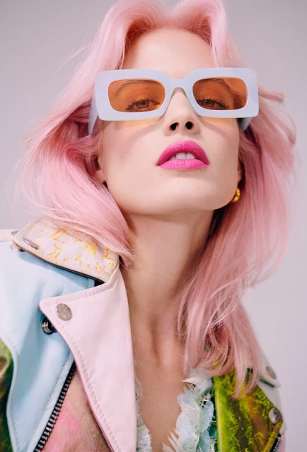 Close-up portrait of a young woman with pink hair wearing a multi-colored jacket and pastel framed sunglasses against a white backdrop.