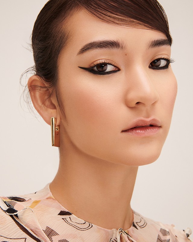 Close-up portrait of a woman with dramatic black winged eyeliner and gold rectangular earrings against a neutral background.