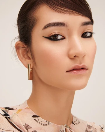 Close-up portrait of a woman with dramatic black winged eyeliner and gold rectangular earrings against a neutral background.