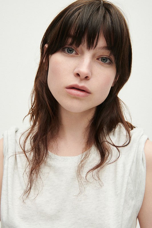 Portrait of a young adult woman with fair skin and dark brown hair in a light gray sleeveless shirt, shot in a photo studio.