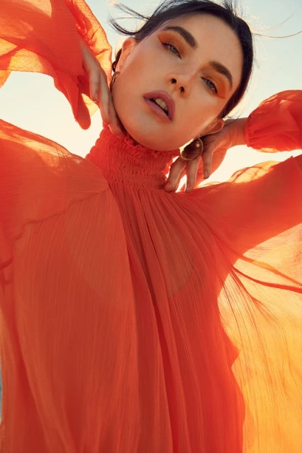 Portrait of a stylish woman in an orange dress with hands near her face, against a bright sky, showcasing fashion and beauty.