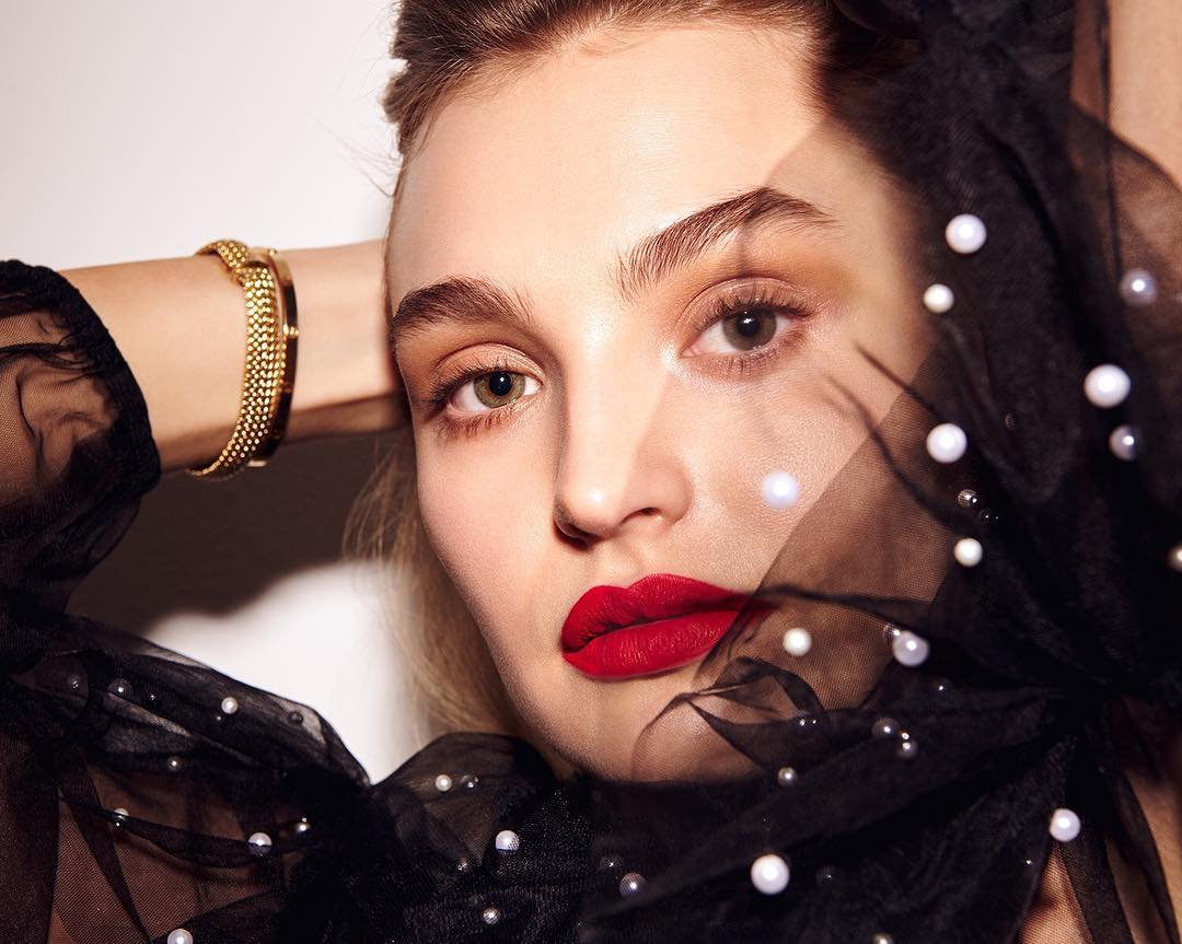 Close-up portrait of a woman with red lipstick wearing a sheer black top with pearls, hands behind head. Beauty and fashion concept.