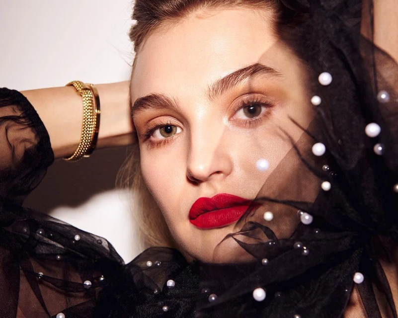 Close-up portrait of a woman with red lipstick wearing a sheer black top with pearls, hands behind head. Beauty and fashion concept.