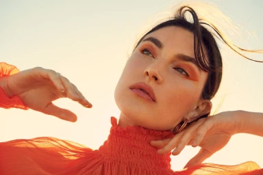 Close-up portrait of a woman wearing an orange ruffled top against a bright sunset sky by photographer Kate Powers.