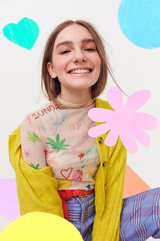 Smiling young woman in fashion portrait with a graphic print mesh top, yellow cardigan, and colorful geometric paper shapes backdrop.