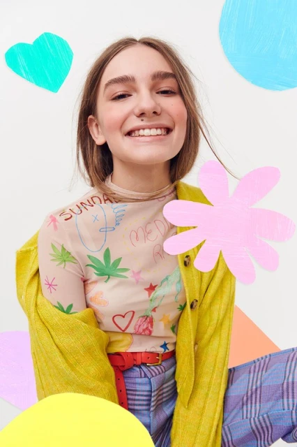 Smiling young woman in fashion portrait with a graphic print mesh top, yellow cardigan, and colorful geometric paper shapes backdrop.
