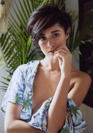 Portrait of a young adult female with a short dark hairstyle, wearing a Hawaiian shirt, and posing indoors with tropical plants behind her.