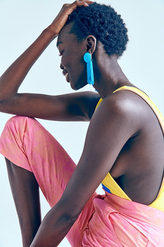 Profile portrait of a Black woman with short hair, wearing blue tassel earrings, a yellow top, and pink pants, smiling in a studio.
