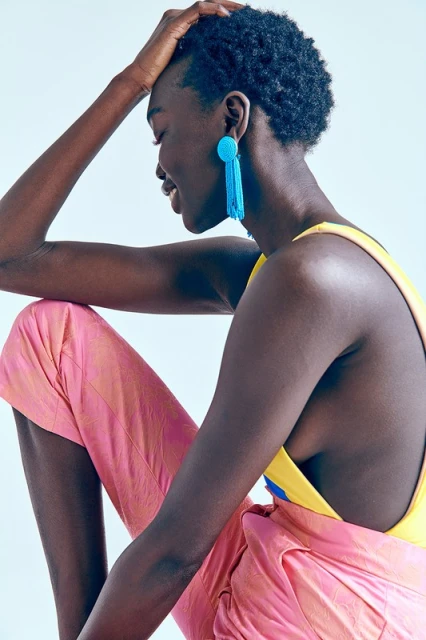 Profile portrait of a Black woman with short hair, wearing blue tassel earrings, a yellow top, and pink pants, smiling in a studio.