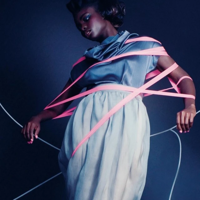 Fashion photo featuring a black female model in a grey satin dress, wrapped in pink ribbons, set against a dark background in a studio.
