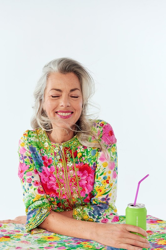 Mature woman with grey hair in floral dress smiles with eyes closed, holding green beverage can with pink straw on white background.