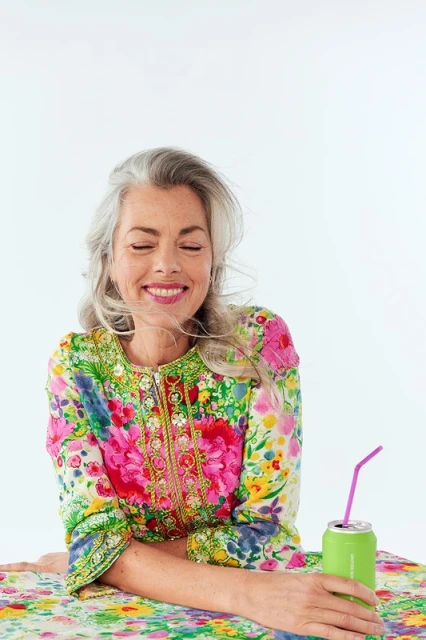 Mature woman with grey hair in floral dress smiles with eyes closed, holding green beverage can with pink straw on white background.