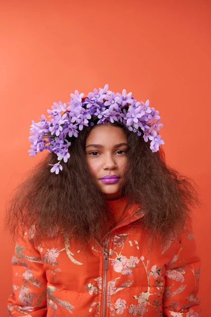 Young woman with purple flower crown and floral print coat posing in front of an orange backdrop in a studio setting.