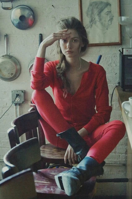 A young adult woman in a red jumpsuit and black boots sits on a wooden chair in an indoor kitchen setting with vintage decor.