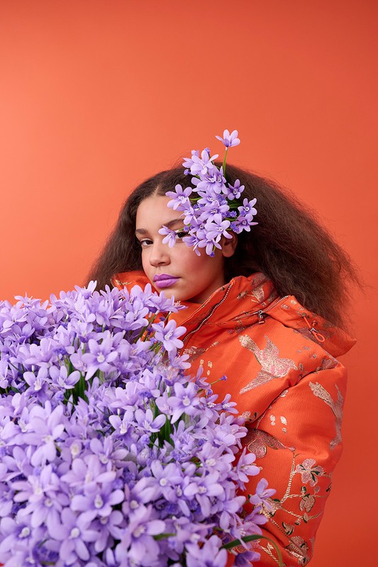 Young woman with purple lipstick and floral headpiece poses behind bouquet of purple flowers against an orange backdrop, studio lighting.