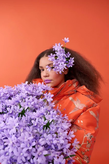 Young woman with purple lipstick and floral headpiece poses behind bouquet of purple flowers against an orange backdrop, studio lighting.