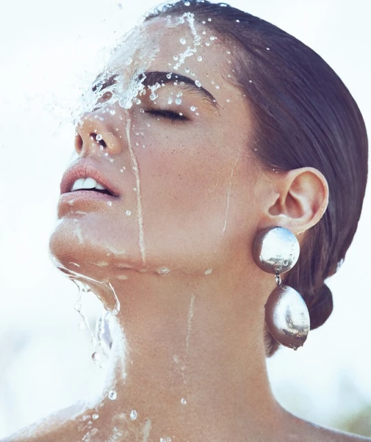 Woman with wet face and eyes closed, water splashes on skin, wearing silver drop earrings, bright light, portrait shot, beauty concept.