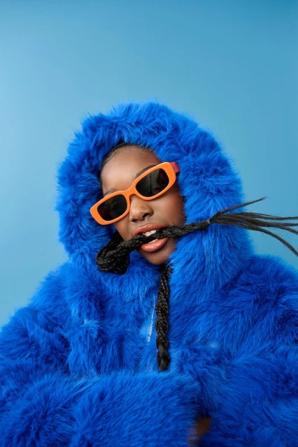 A woman with dark skin and braids poses in a bright blue furry hooded coat and orange sunglasses against a solid blue backdrop.