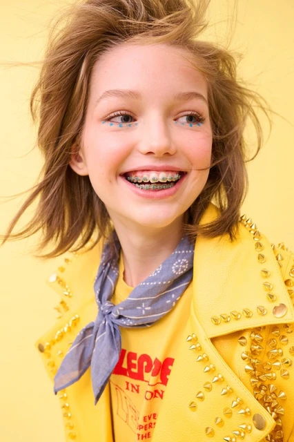 Smiling child with braces and star-shaped makeup wearing a studded yellow jacket, captured in a vibrant studio portrait on yellow backdrop.