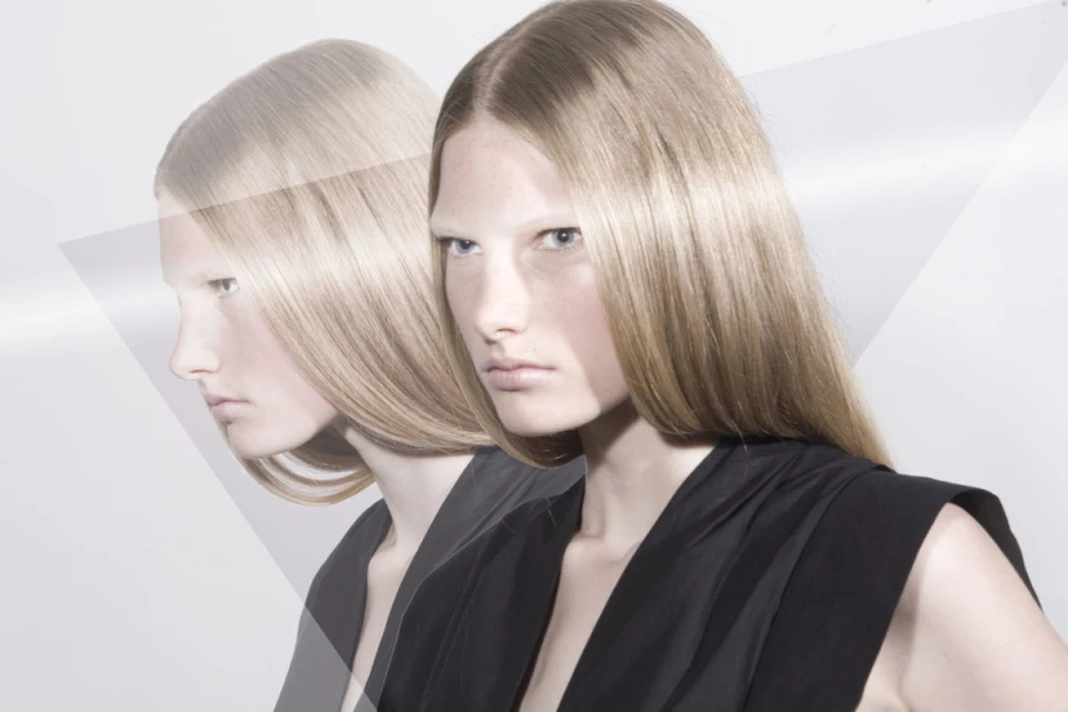 Blonde woman with pale eyebrows wearing a black top against a white background, with a ghosted profile visible.
