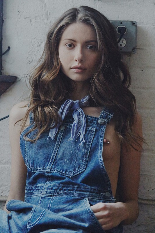 Brunette woman in denim overalls and scarf poses against textured wall in a medium shot, showcasing fashion and simplicity.