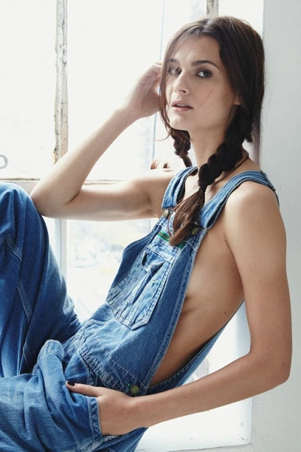 Brunette woman in blue denim overalls, posing in front of a window with natural lighting, wearing pigtails, and looking at the camera.