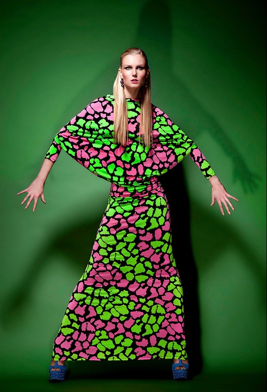 Woman in a brightly colored dress strikes a pose in front of a green screen. The long-sleeved dress has a pink and green pattern.