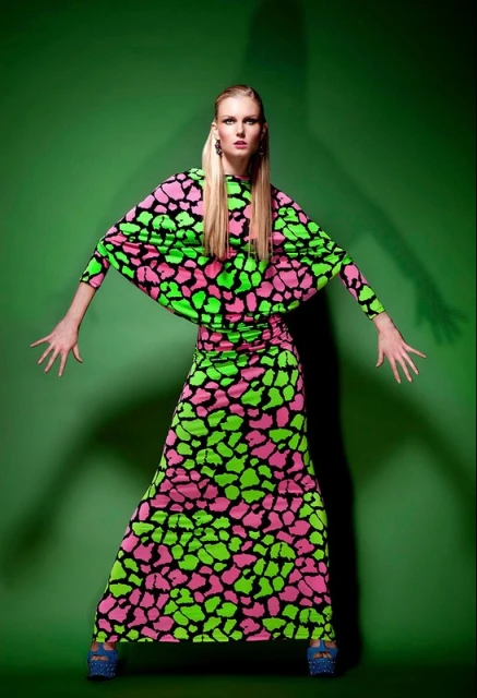 Woman in a brightly colored dress strikes a pose in front of a green screen. The long-sleeved dress has a pink and green pattern.