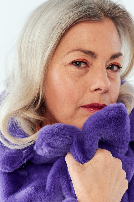 Close-up portrait of a mature woman with gray hair wearing a vibrant purple faux fur jacket in a photo studio setting.