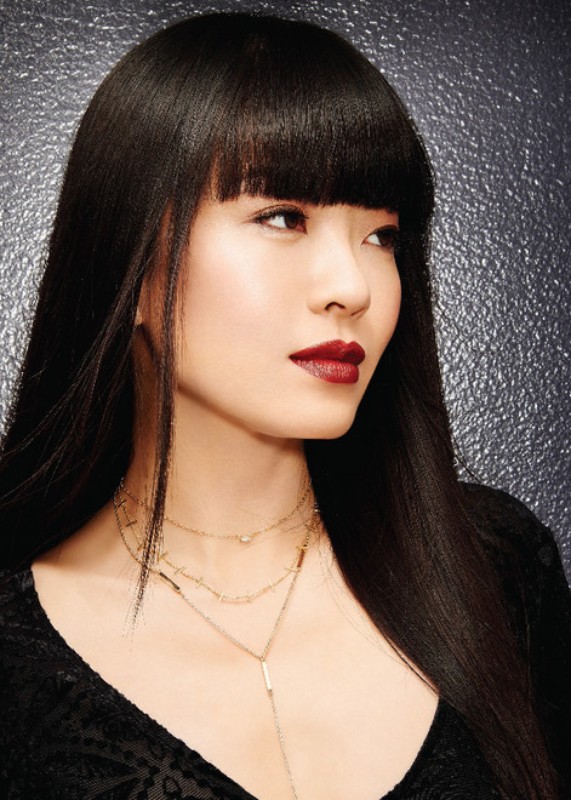 Portrait of a striking Asian woman with sleek dark hair, dark lipstick, and delicate gold necklaces against a textured backdrop.