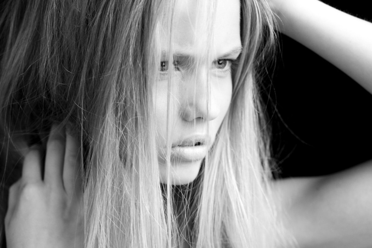 Close-up monochrome portrait of a woman with long hair framing her face and hands raised near her head against a dark background.