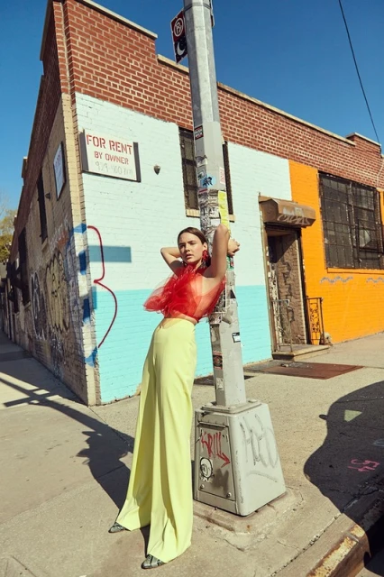 Fashion shot of a young adult woman outdoors with long pants and a sheer red top, leaning against a street pole on a sunny day.