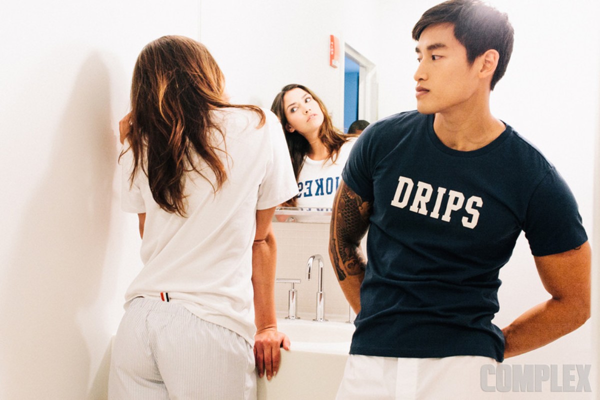 Three people stand in a brightly lit bathroom: a woman facing a mirror, a woman with her back to the camera, and an Asian man.