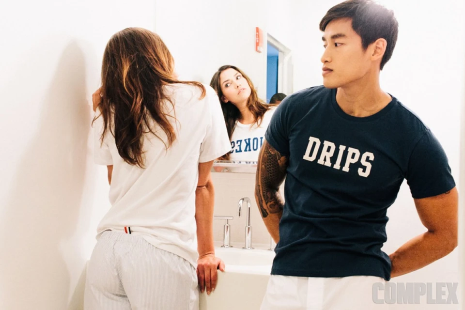 Three people stand in a brightly lit bathroom: a woman facing a mirror, a woman with her back to the camera, and an Asian man.