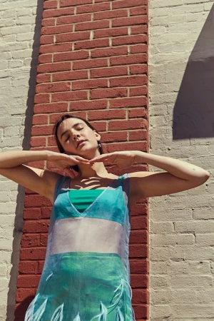 A young woman with her eyes closed wearing a sheer turquoise dress against a brick wall in Los Angeles.