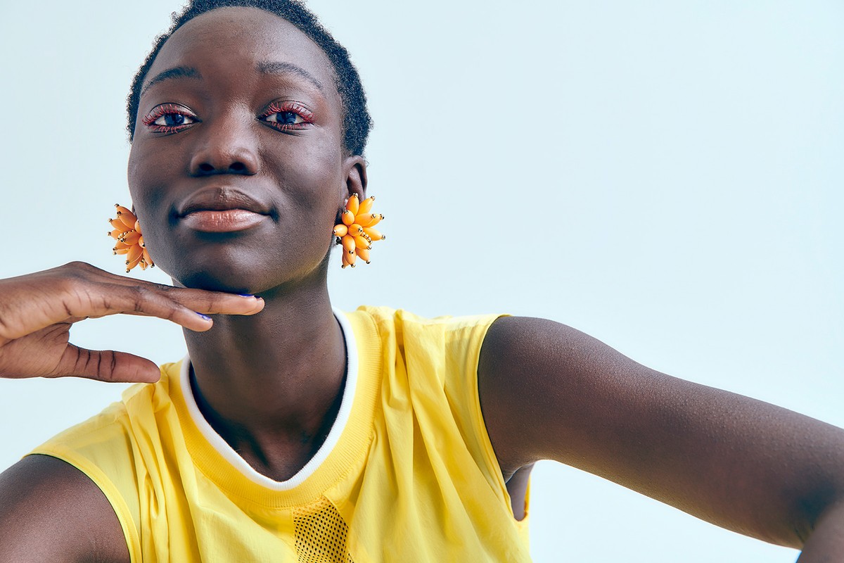 Close-up portrait of a young Black woman with orange floral earrings and a yellow athletic top, posing in front of a white backdrop.