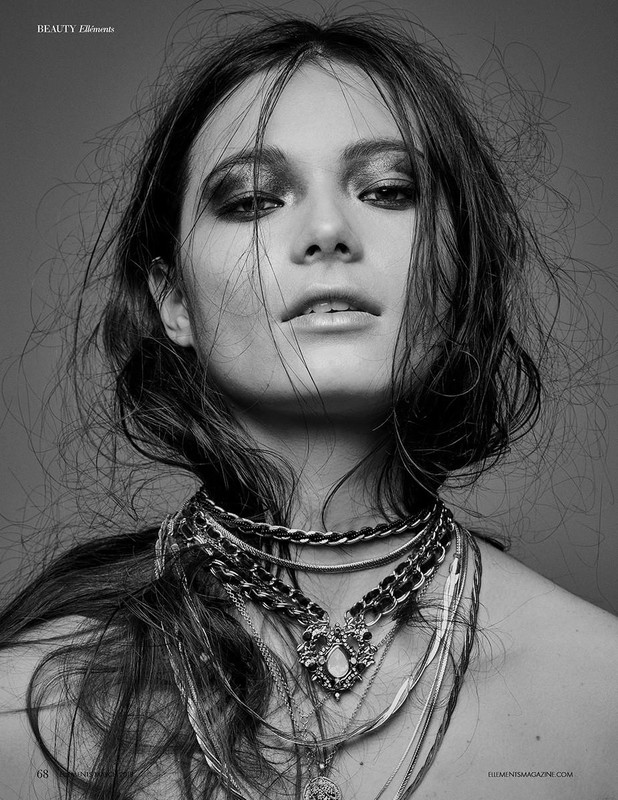 Close-up monochrome portrait of a woman with smoky eye makeup and layered necklaces against a grey background.
