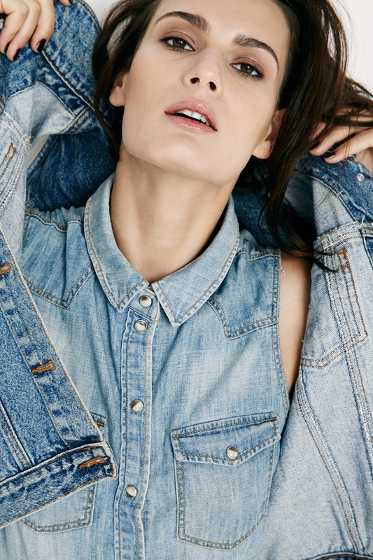 Close-up portrait of a woman wearing a light blue denim vest and holding a matching denim jacket against a plain background