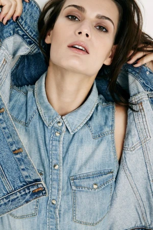 Close-up portrait of a woman wearing a light blue denim vest and holding a matching denim jacket against a plain background