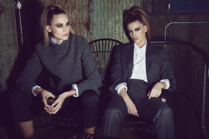 Two women in formal attire are seated indoors, one in a suit and the other in a sweater, posing for a fashion editorial style portrait.
