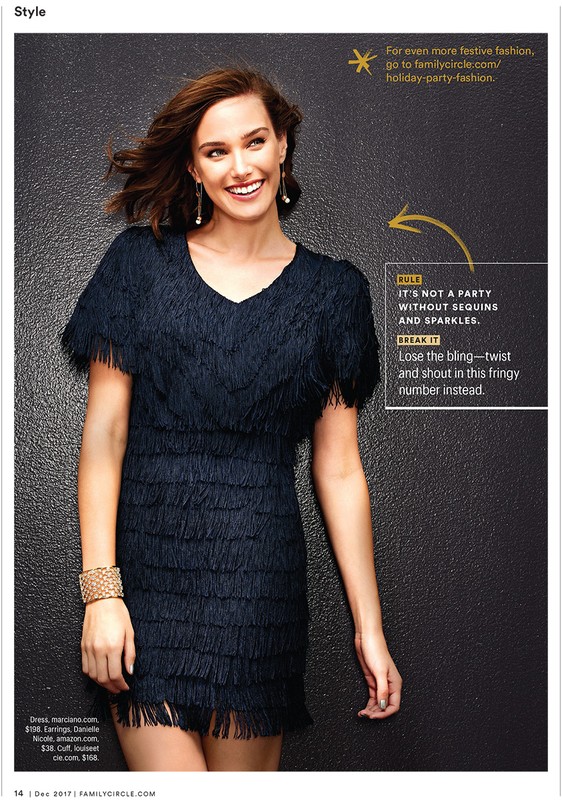 Smiling woman in a fringed black dress and gold bracelet poses against a dark, textured background in a fashion editorial shot.