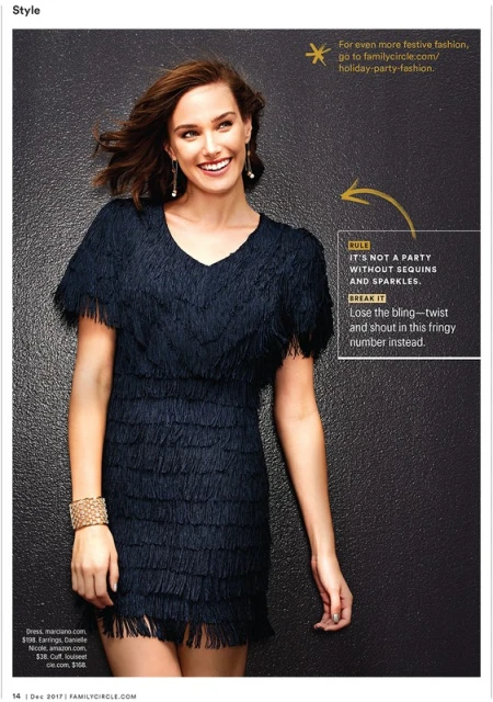 Smiling woman in a fringed black dress and gold bracelet poses against a dark, textured background in a fashion editorial shot.