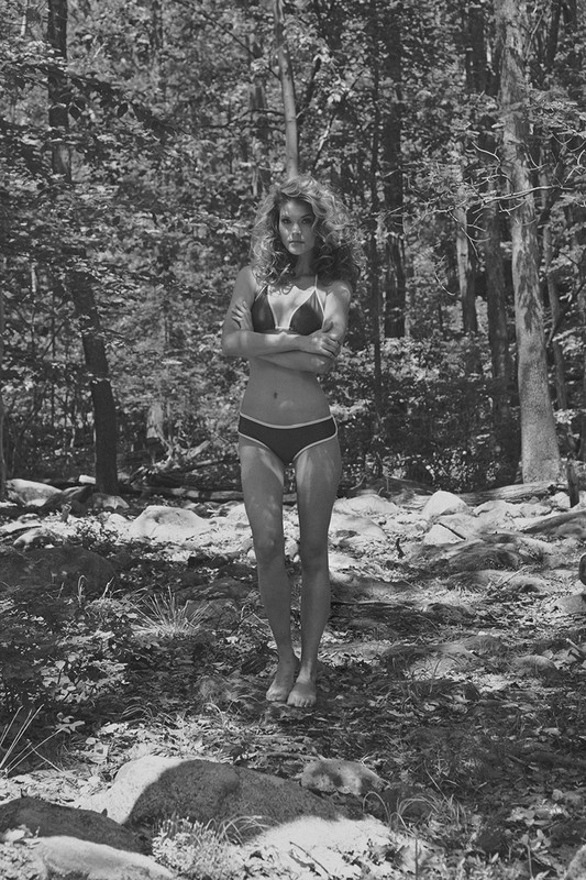A woman stands in a bikini in a grayscale photo, set in a densely wooded forest landscape, with sun dappling through the leaves.