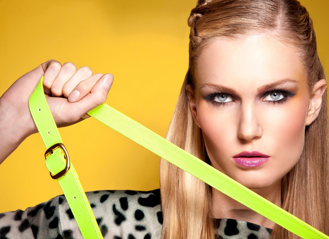 Close-up portrait of a blonde woman with dramatic makeup holding a neon green belt against a bright yellow background, showcasing fashion and beauty.
