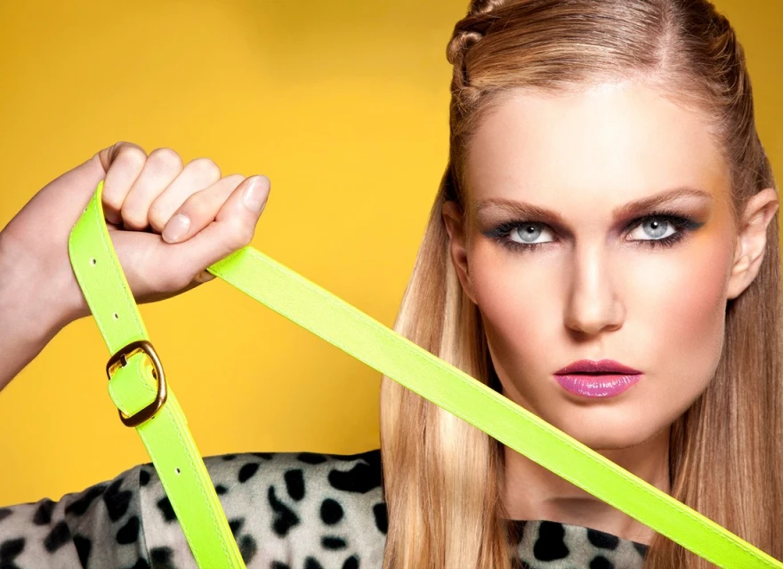 Close-up portrait of a blonde woman with dramatic makeup holding a neon green belt against a bright yellow background, showcasing fashion and beauty.