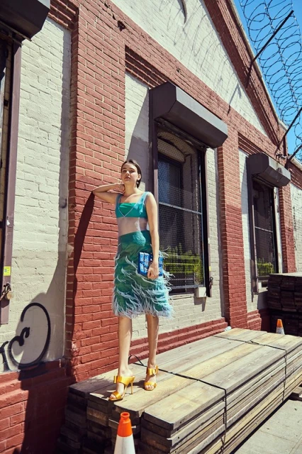 A woman in a turquoise top and green feathered skirt poses on a wooden platform with a brick building and blue sky in the background.