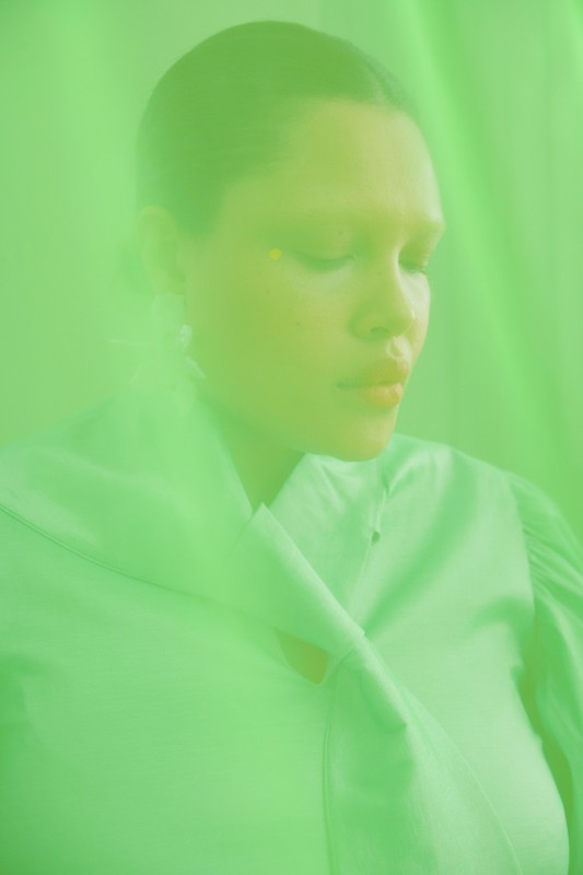 Close-up portrait of a woman with closed eyes, luminous skin, and bright yellow makeup in a monochromatic green environment with soft lighting.